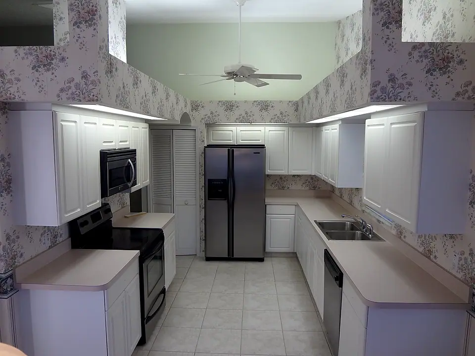 Dated galley kitchen with white laminate cabinets and floral wallpaper, before the remodel